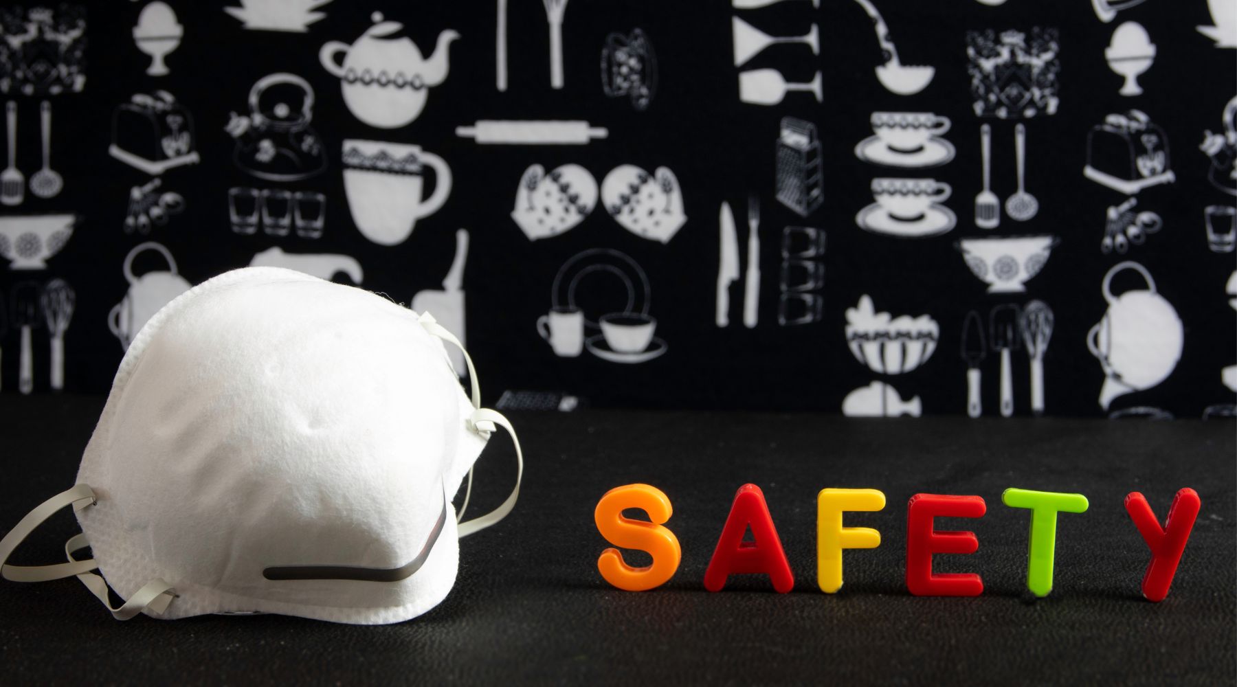 A white face mask sits next to colorful letters spelling SAFETY on a dark surface, reminding everyone to prioritize health even during the holiday season.