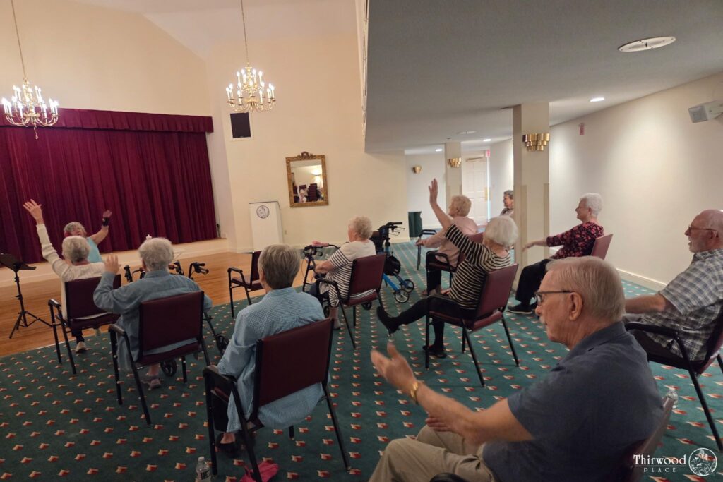 A group of seniors sitting in chairs, raising their hands, faces a stage in a community room during a lively scholarship awards ceremony.