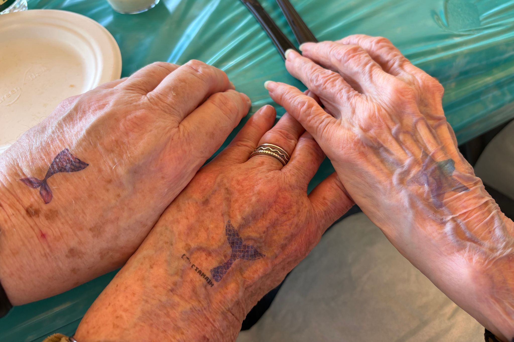 Three elderly hands with mermaid tail temporary tattoos rest on a turquoise tablecloth, capturing the carefree spirit of summer.