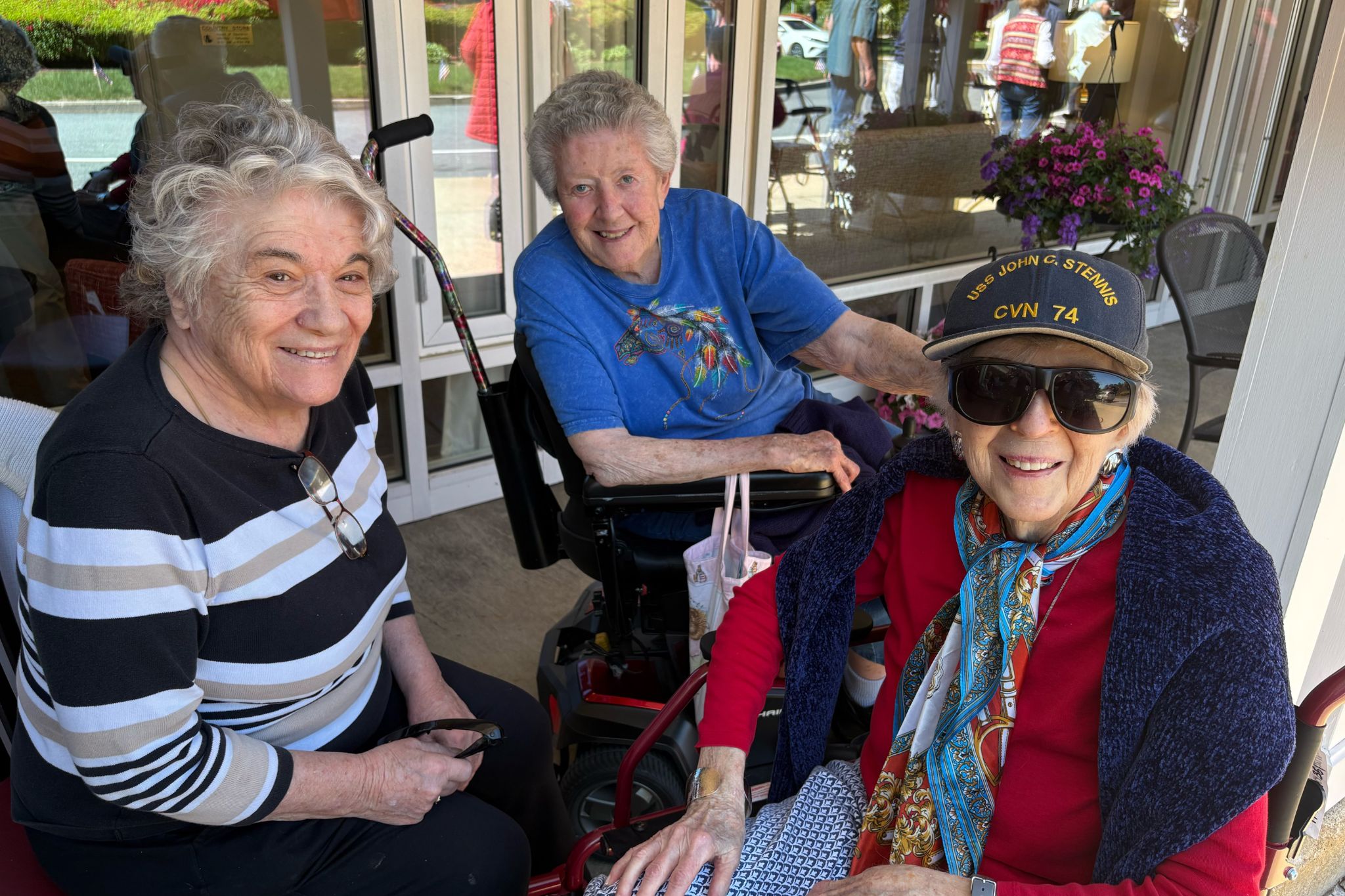 Three smiling older women sitting together outdoors, one wearing sunglasses and a Navy hat, enjoying a sunny summer day.