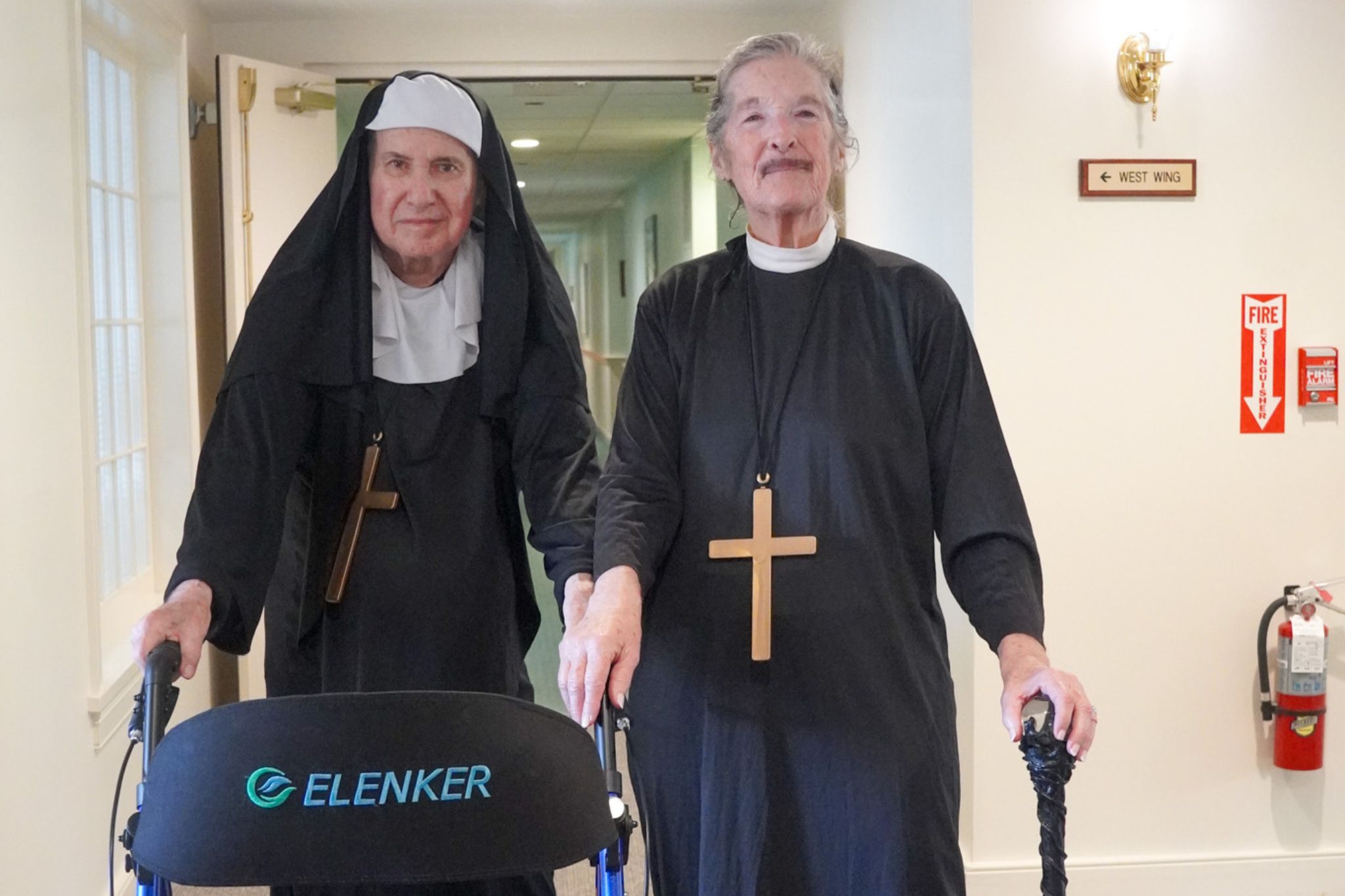 Two elderly people dressed as nuns, each with a large cross necklace, stand in a hallway with mobility aids, sharing stories about their years of service and scholarship.