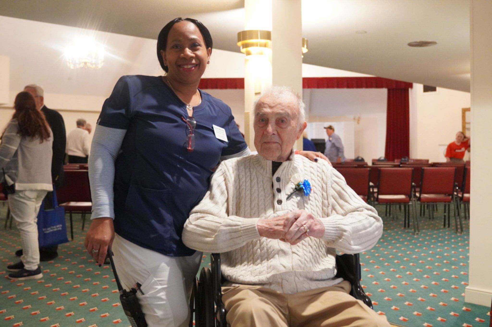 A nurse with a scholarship smiles beside an elderly man in a wheelchair in a room with red chairs and green carpet.
