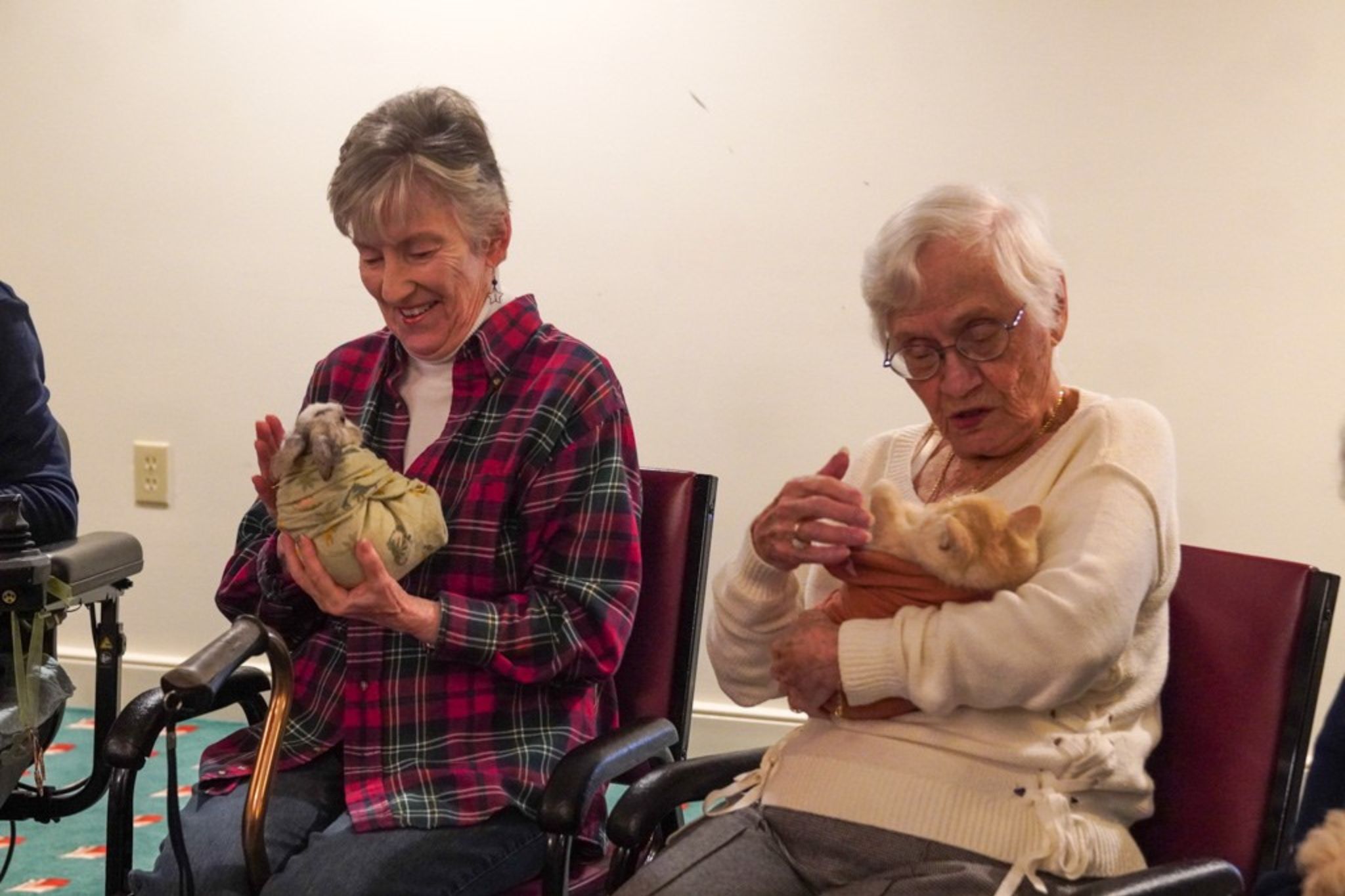 Two elderly women sitting and smiling, each holding a small animal wrapped in a blanket on their laps, celebrate a lifelong friendship sparked by a shared scholarship in their youth.
