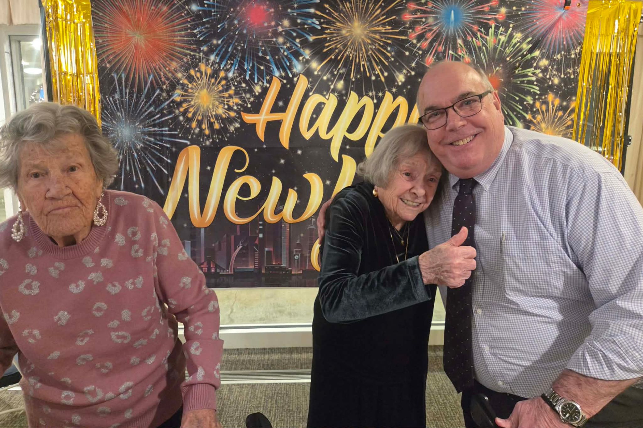 Three people pose and smile in front of a festive Happy New Year backdrop with fireworks graphics, creating the perfect moment for a photo essay capturing summer celebrations.