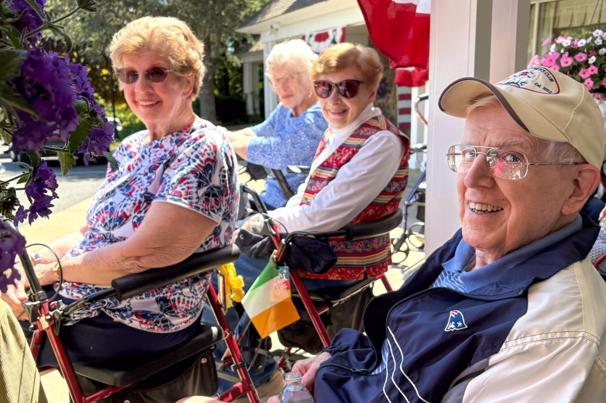 Four smiling older adults sit outdoors with walkers, enjoying a sunny summer day surrounded by flowers.