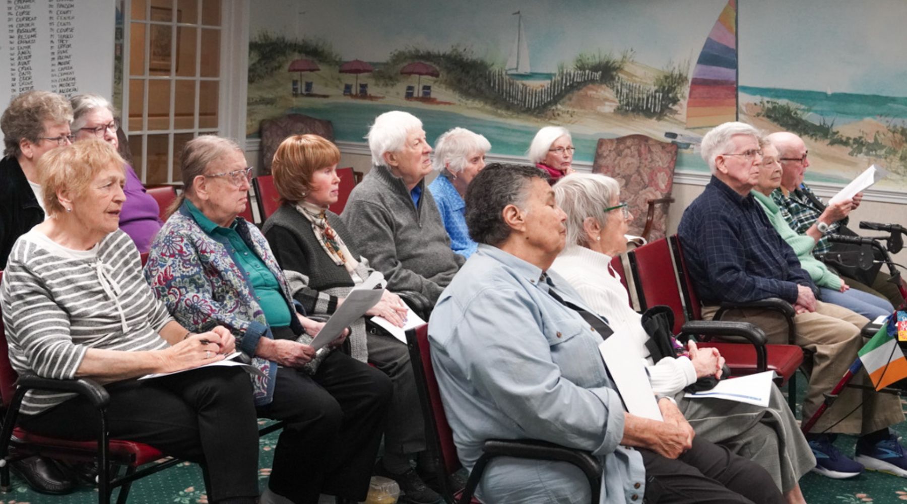 A group of elderly people sit in chairs, holding papers, facing forward in a room with a beach mural decorated with a seascape.