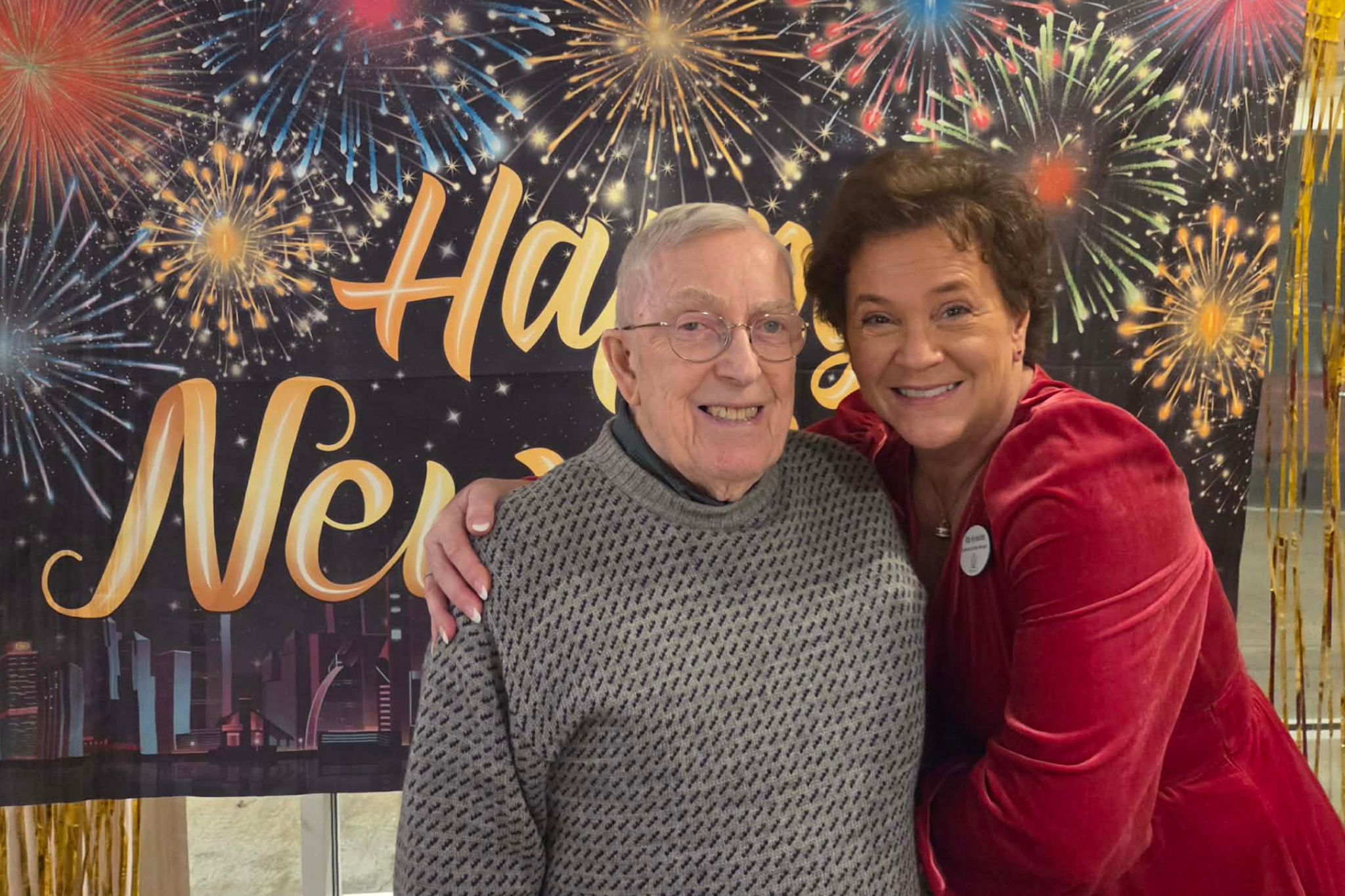 Older man and woman smiling and hugging in front of a Happy New Year fireworks backdrop, capturing a joyful moment perfect for a photo essay—even as summer memories linger in their warm embrace.