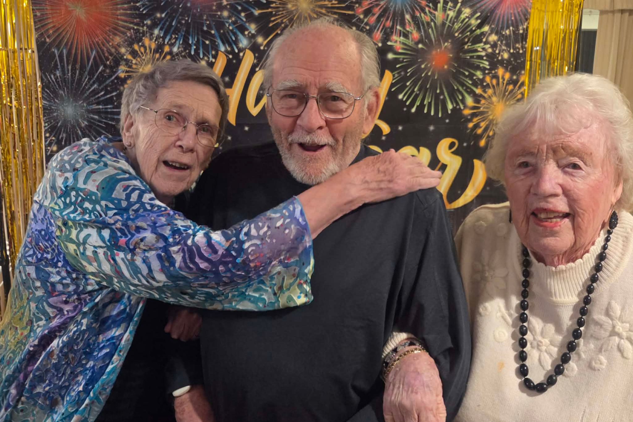 Three elderly people smiling and posing together at a summer celebration with a fireworks-themed backdrop, perfect for a joyful photo essay.