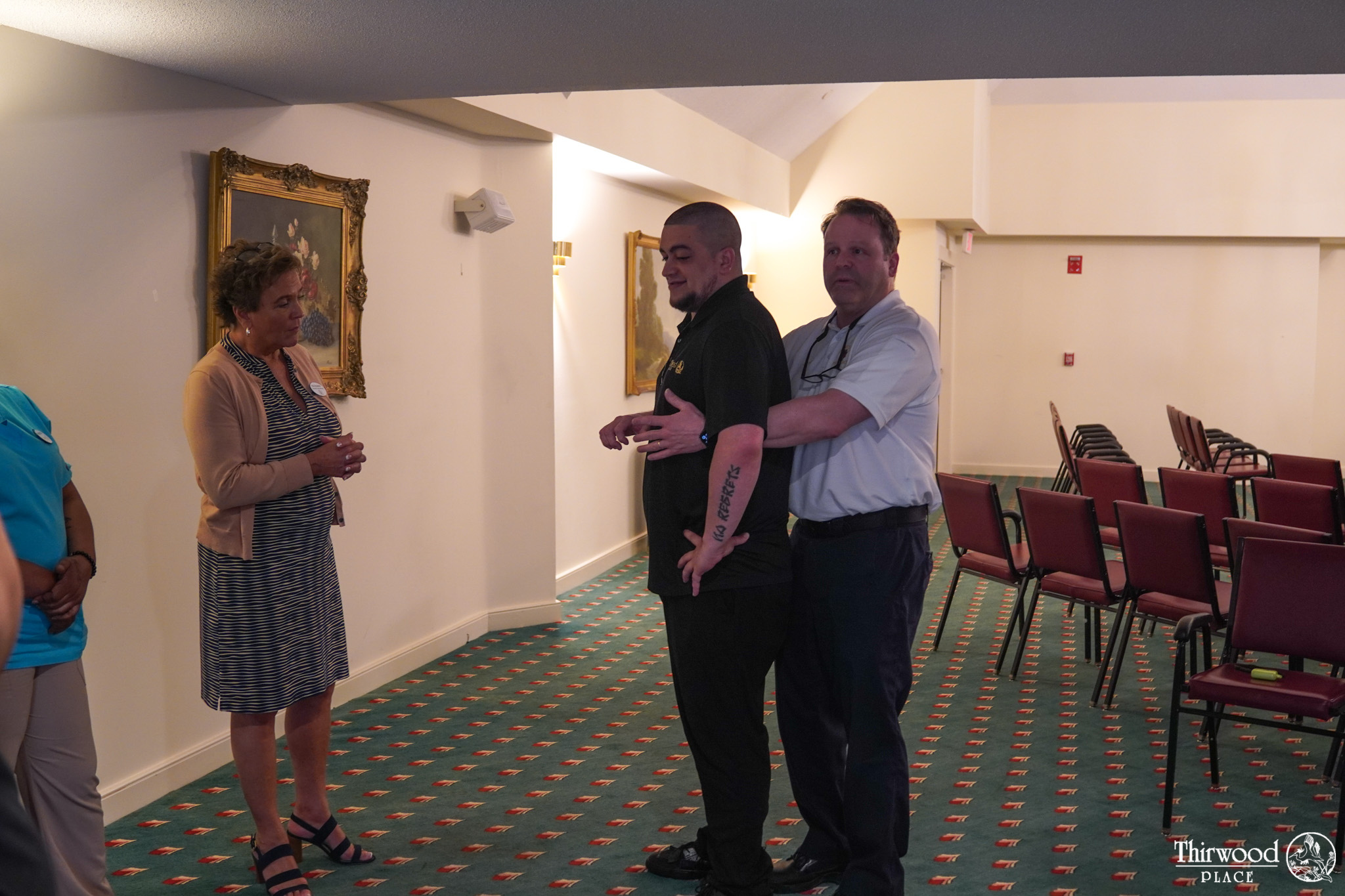 Two men demonstrate the Heimlich maneuver, possibly in response to a food emergency, as two women watch in a carpeted room with chairs and a painting.