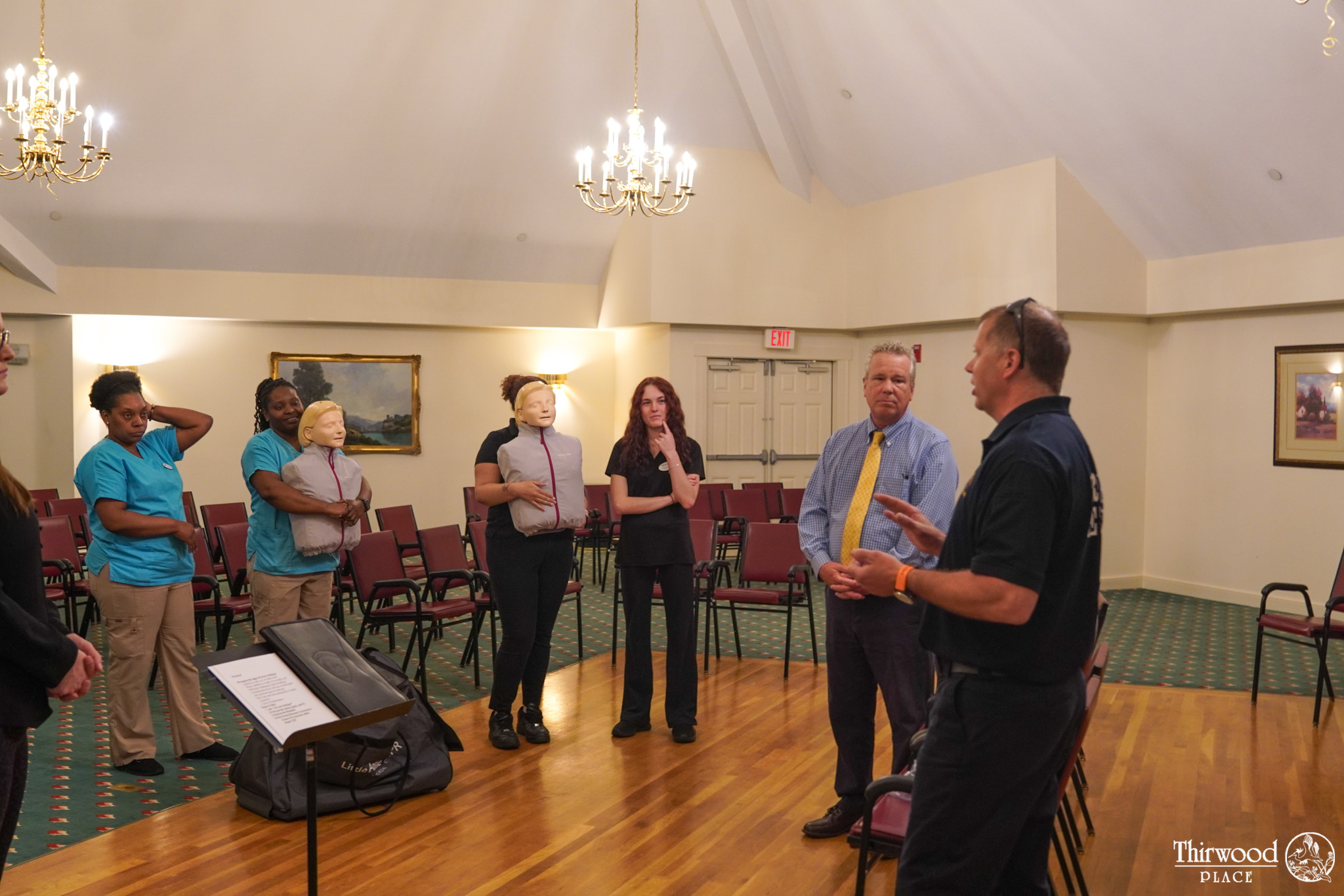 A group listens to a speaker in a room; some people hold training mannequins with CPR faces as they discuss cold and flu prevention.