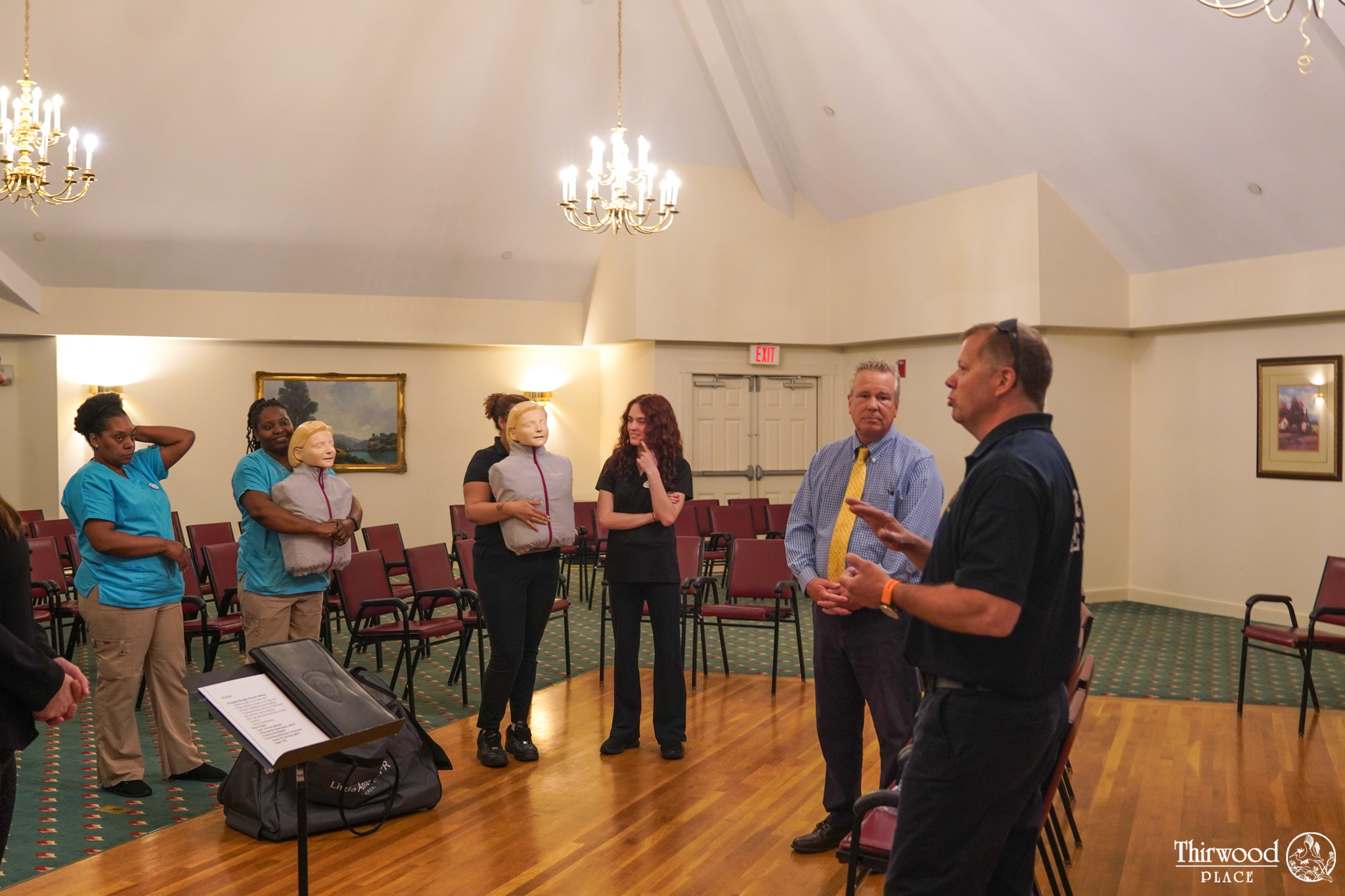 People in a room listen to a man speaking; two hold medical training mannequins, likely discussing cold and flu scenarios, among empty chairs.