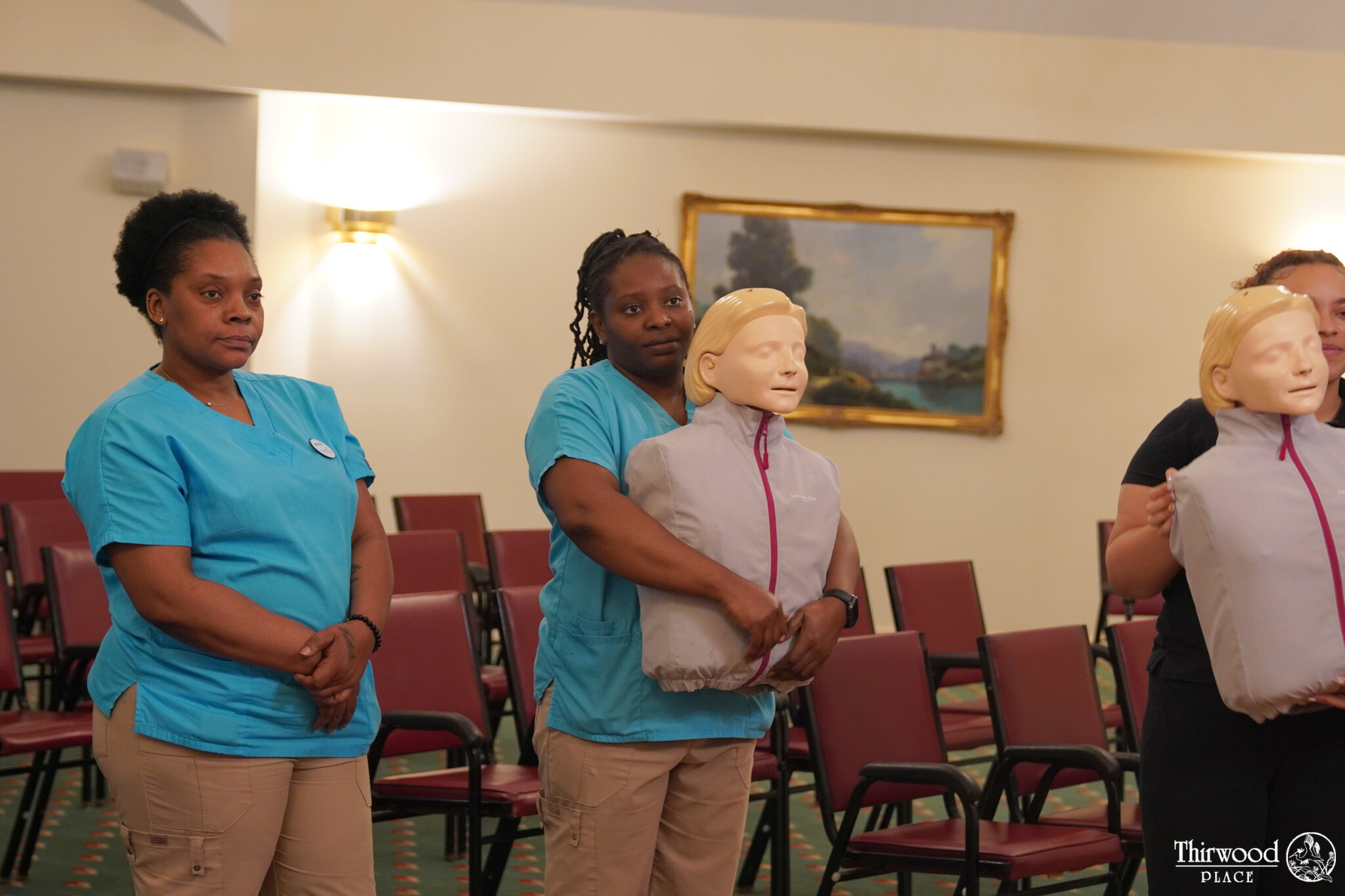 Three people in scrubs hold CPR training mannequins in an empty room with chairs and a painting on the wall, preparing for a session on emergency care, including tips on cold and flu prevention.