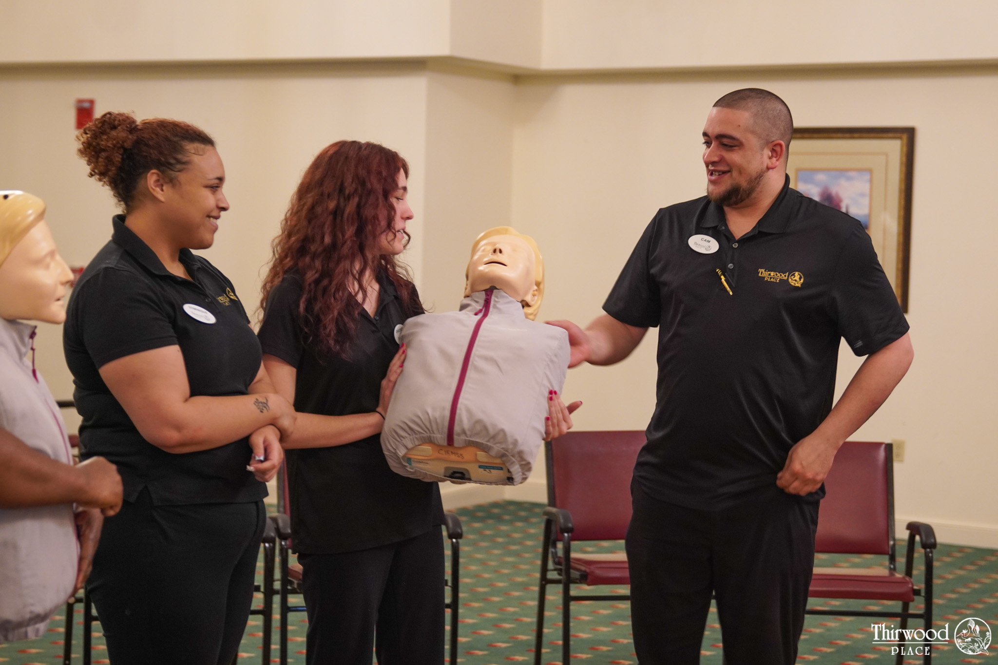 Three people smile while holding a CPR training dummy during a class in a room with chairs and framed art, learning lifesaving skills to help others in emergencies like cold and flu seasons.