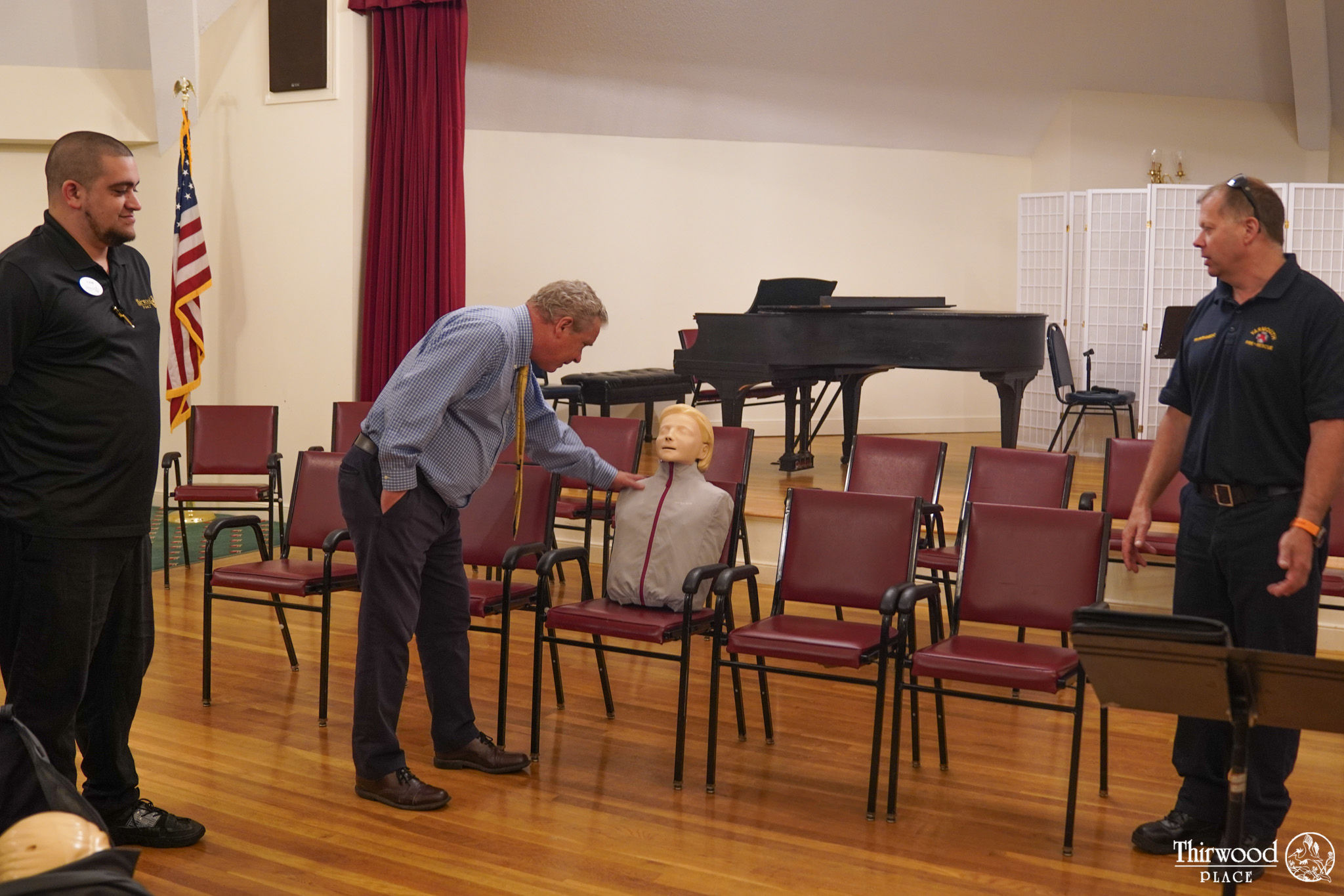 A man demonstrates CPR on a practice dummy while two others observe in a room with chairs, a piano, and posters about cold and flu prevention.