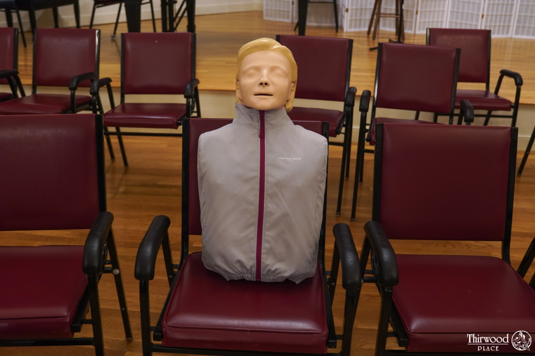 A CPR training mannequin in a jacket, perfect for cold and flu season demos, sits on a chair among rows of empty red chairs.