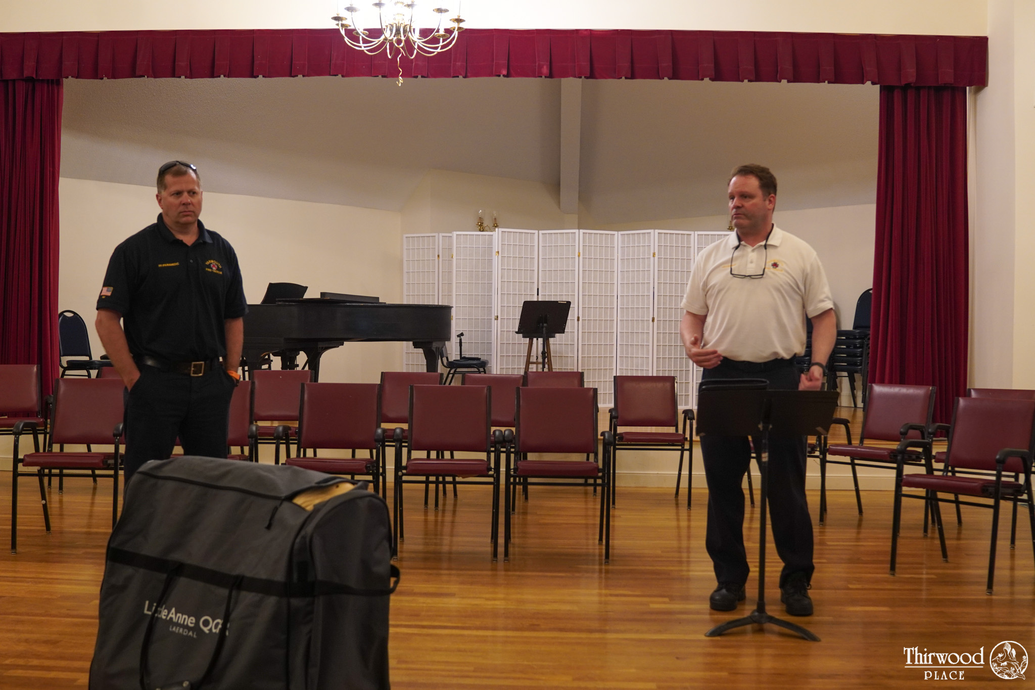 Two men stand and face forward in a room with chairs, a piano, and a red-curtained stage in the background, perhaps discussing food or sharing tips on staying healthy during cold and flu season.