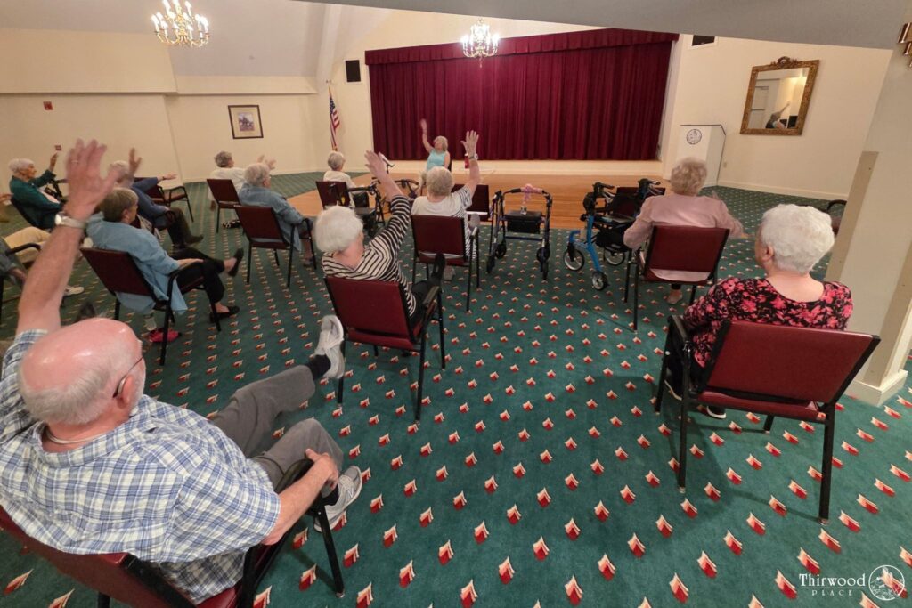Seniors seated in a room raise their hands, facing a stage with a red curtain at a community event celebrating a scholarship award.