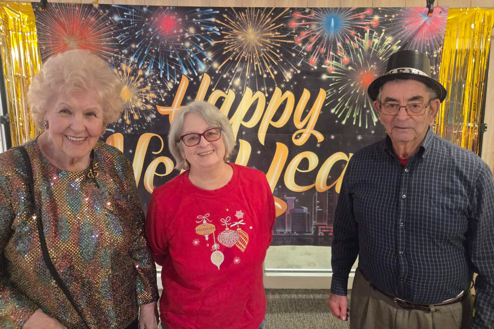Three older adults smiling in front of a Happy New Year banner decorated with fireworks, captured as part of a joyful photo essay.