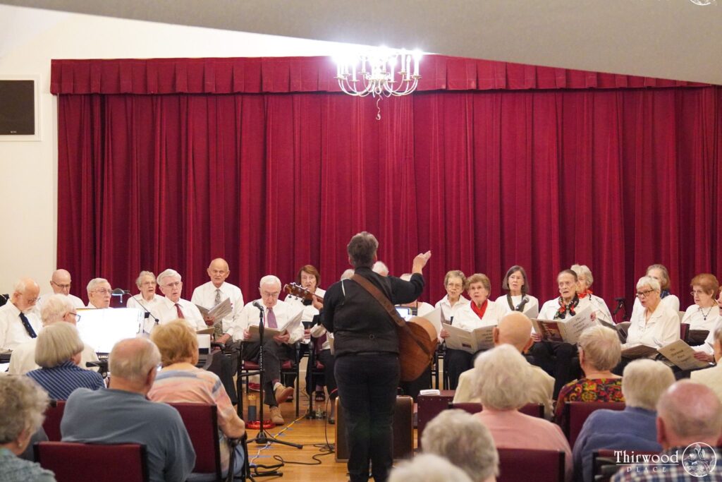 A choir performs on stage with a conductor, facing an audience in a room with red curtains, celebrating talented voices brought together through a scholarship program.