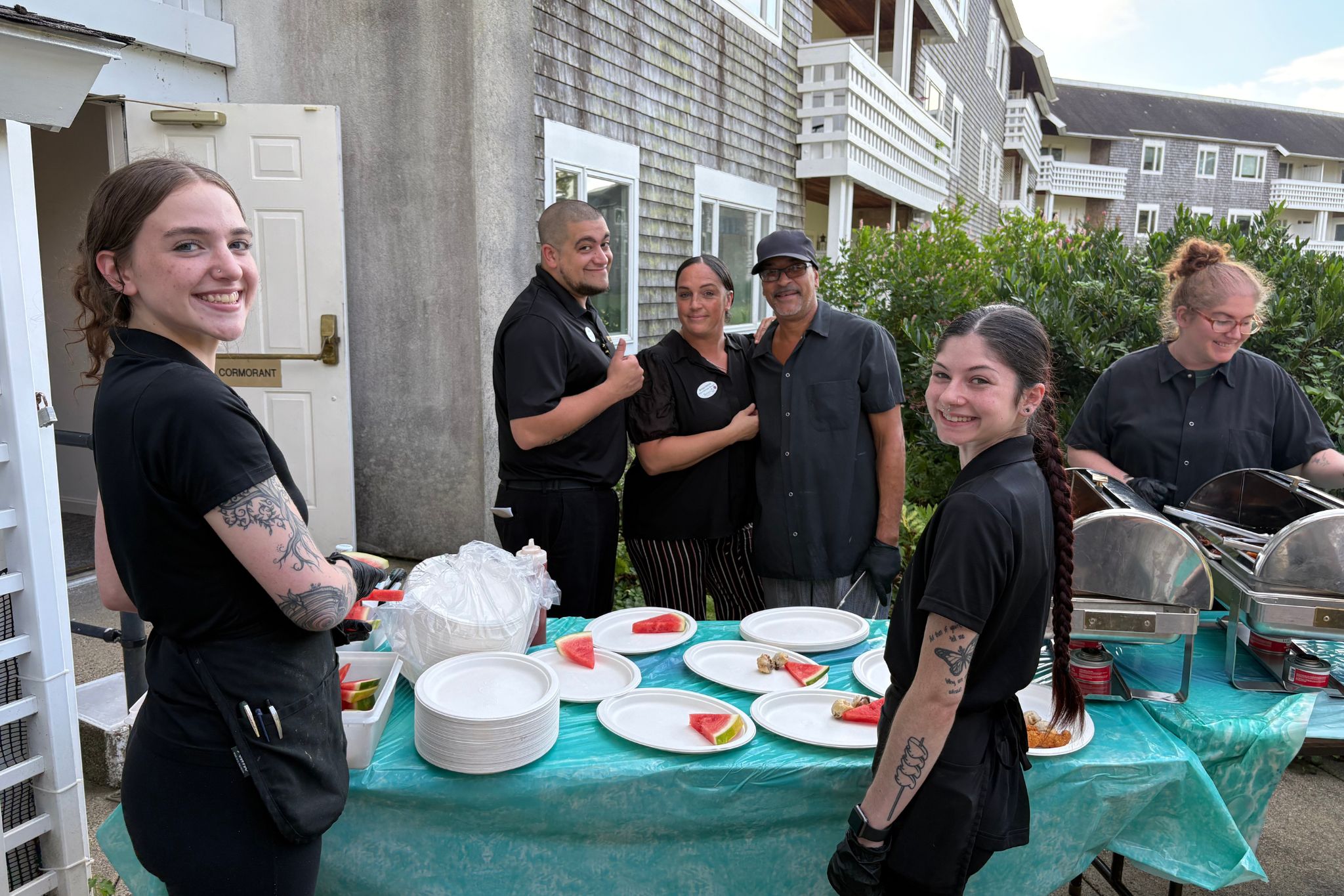 Five smiling people in black uniforms serve food and watermelon slices at an outdoor summer event table.