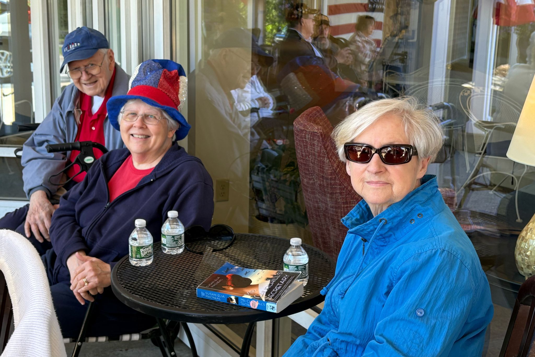 Three older adults sit by a table outside, smiling and enjoying the summer; one woman wears a festive hat, another sunglasses.