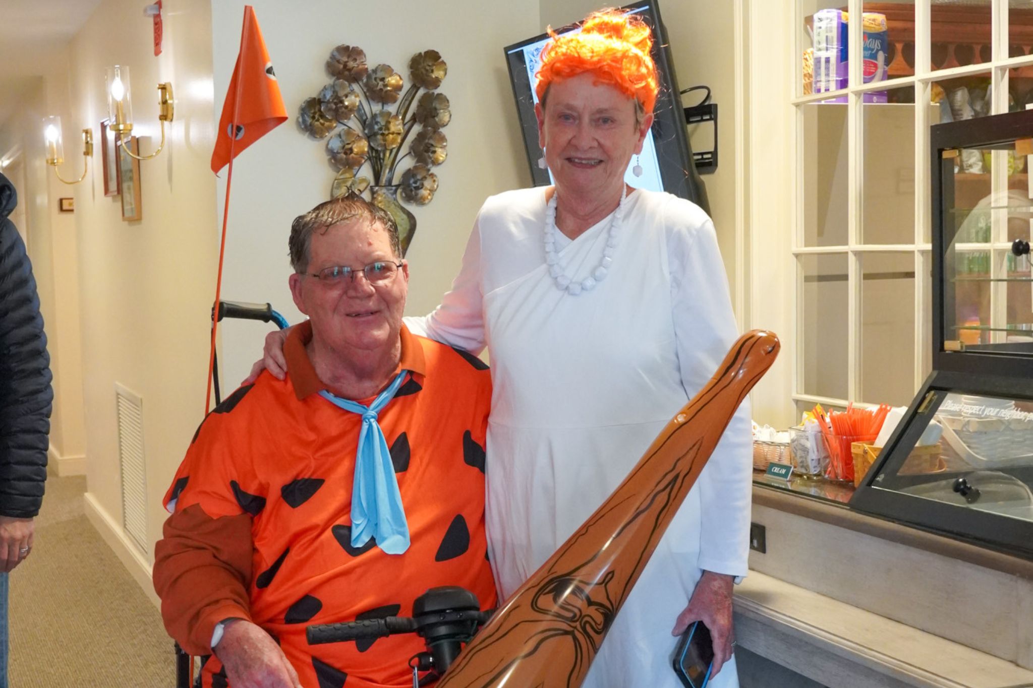 Two adults in Flintstones costumes, one seated as Fred and one standing as Wilma, smiling indoors, celebrate their scholarship win with prehistoric flair.