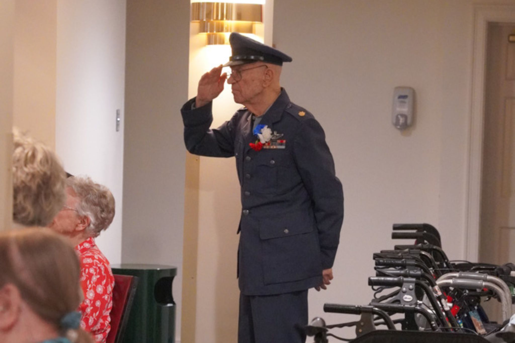 An elderly man in a military uniform salutes while standing indoors near seated people and walkers, celebrating his service and the scholarship opportunities it has inspired for future generations.