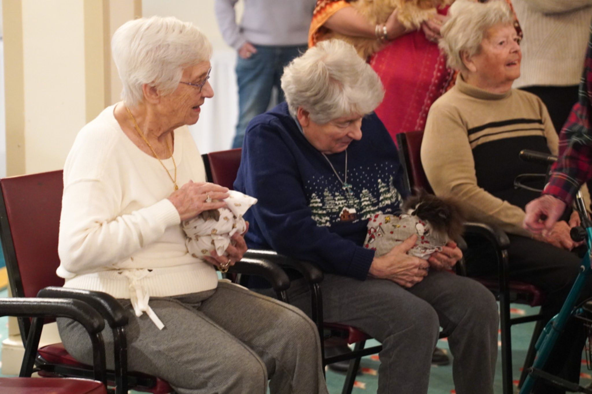 Three elderly women sit with gifts on their laps, one proudly holding a small dog, in a festive indoor setting—gathered to celebrate a special scholarship announcement.