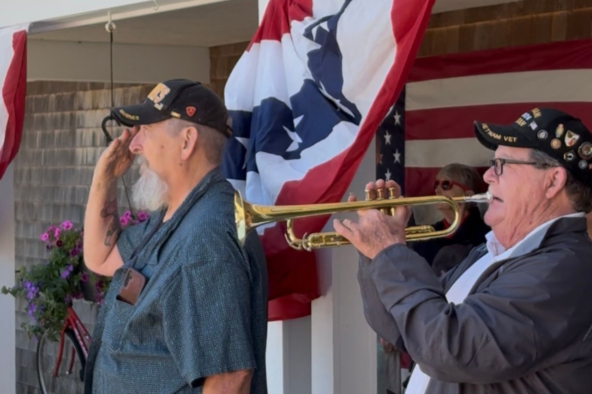 Two older men, one saluting and one playing trumpet, stand by patriotic banners and flags on a bright summer day.