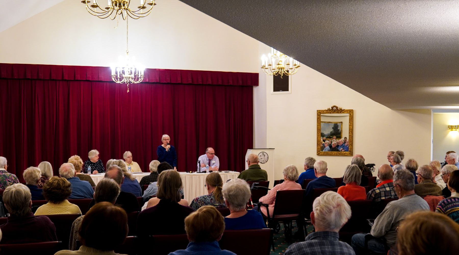 A group of people listen to a panel of speakers at a meeting in a room with red curtains, chandeliers, and subtle animal motifs adorning the decor.