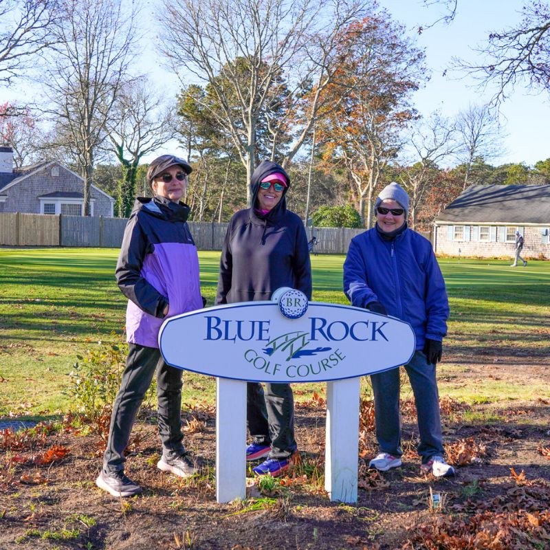 Three people in jackets and sunglasses stand by a Blue Rock Golf Course sign on a sunny Cape Cod day.