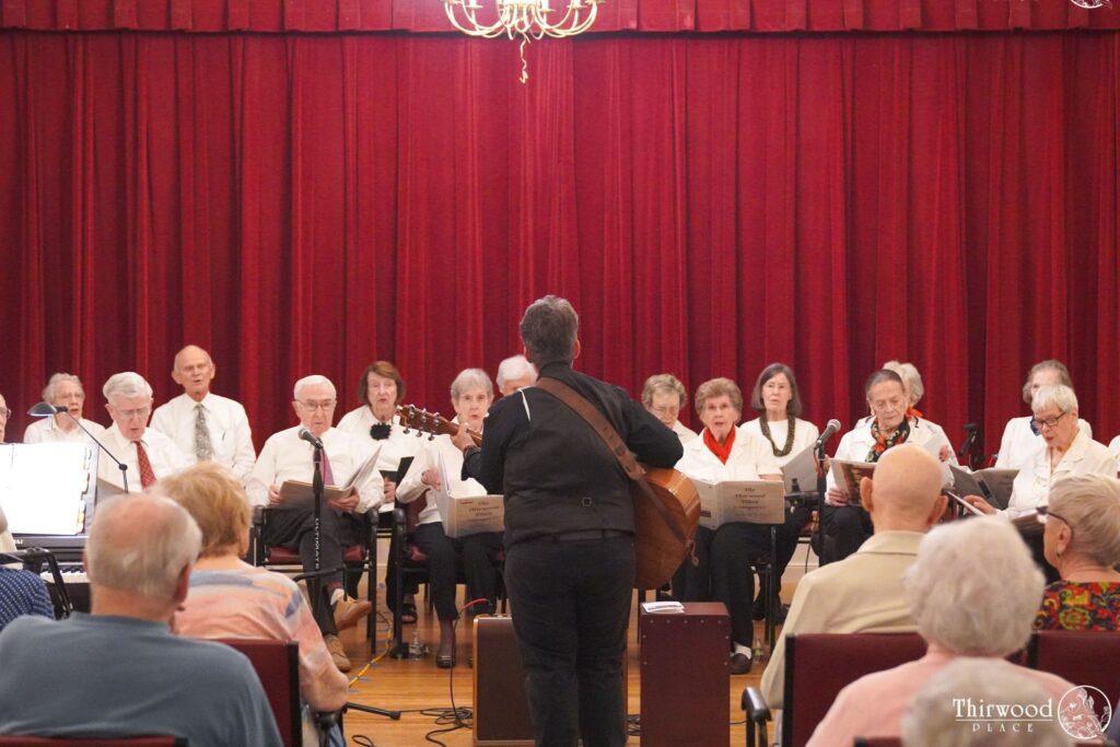 A choir of older adults, brought together through a scholarship program, performs on stage with a conductor playing guitar, facing an audience.