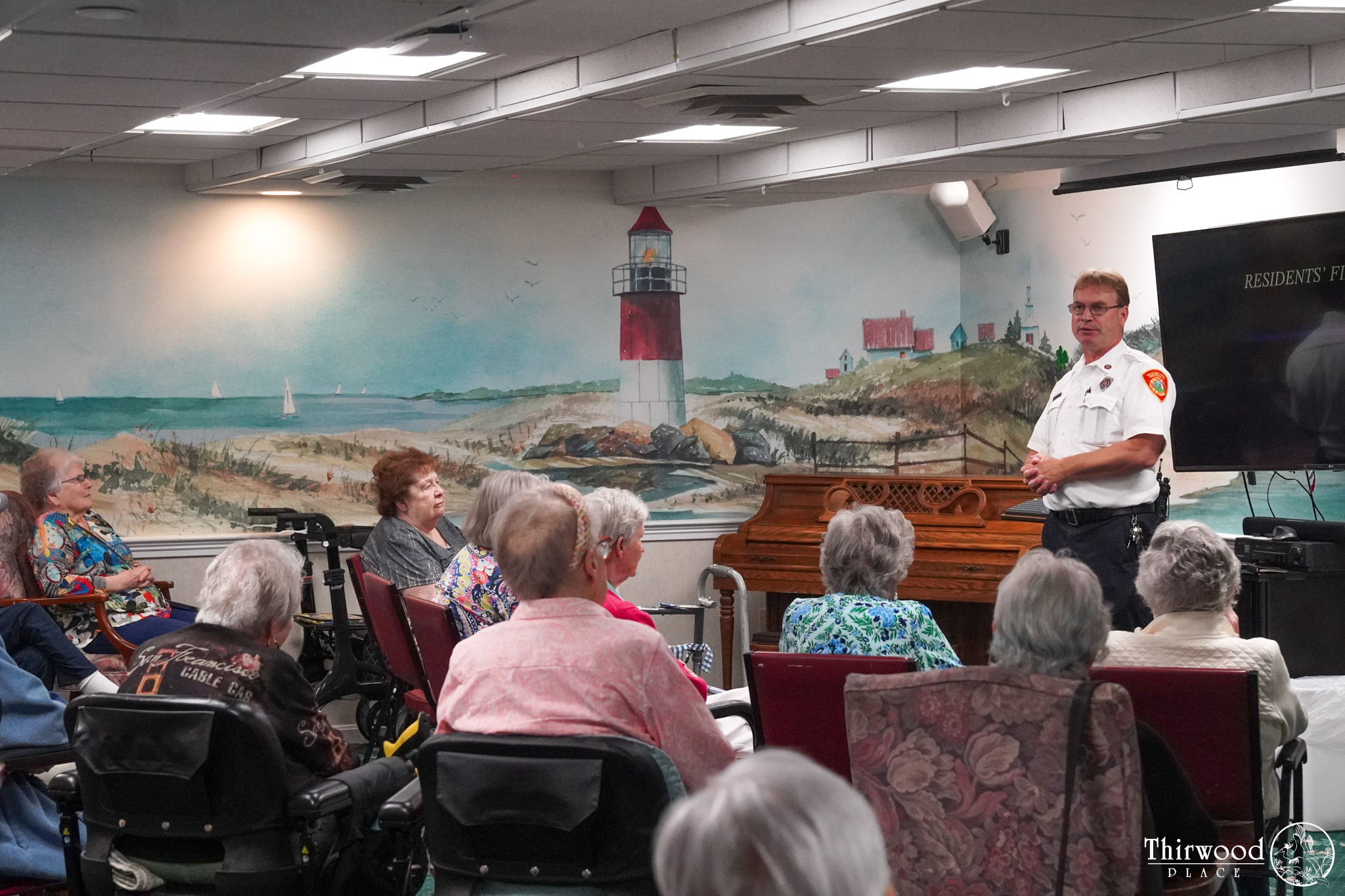 A firefighter speaks to a group of seniors seated in a room with a lighthouse mural, sharing tips on how to prevent cold and flu or respond if someone starts to choke.