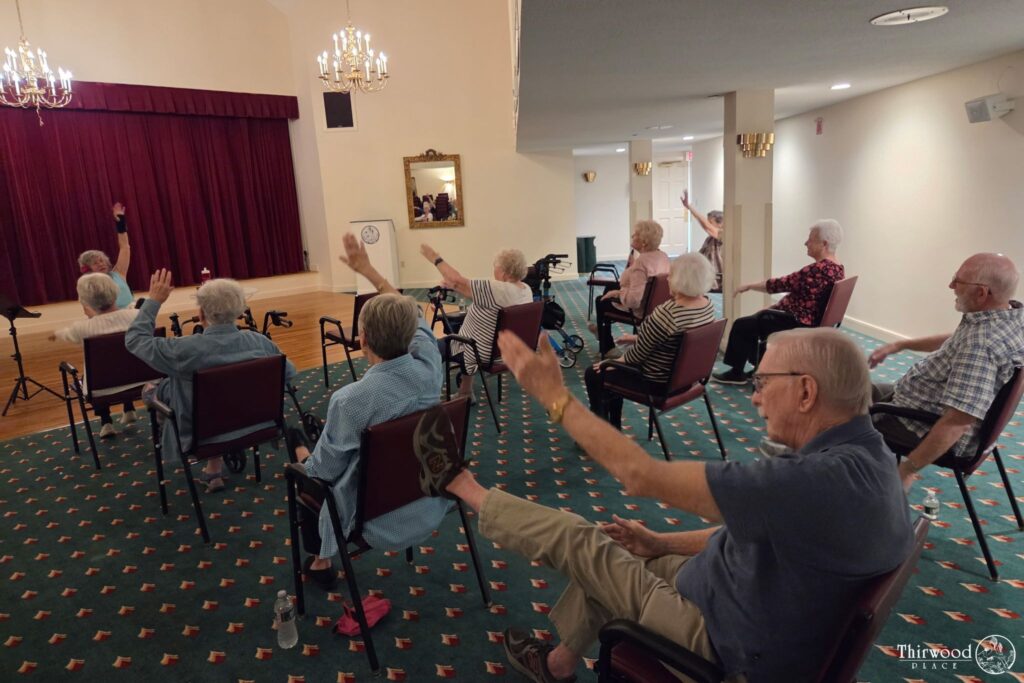 Seniors seated in chairs participate in a group exercise, raising arms and legs in a carpeted room, while discussing scholarship opportunities for continuing education.