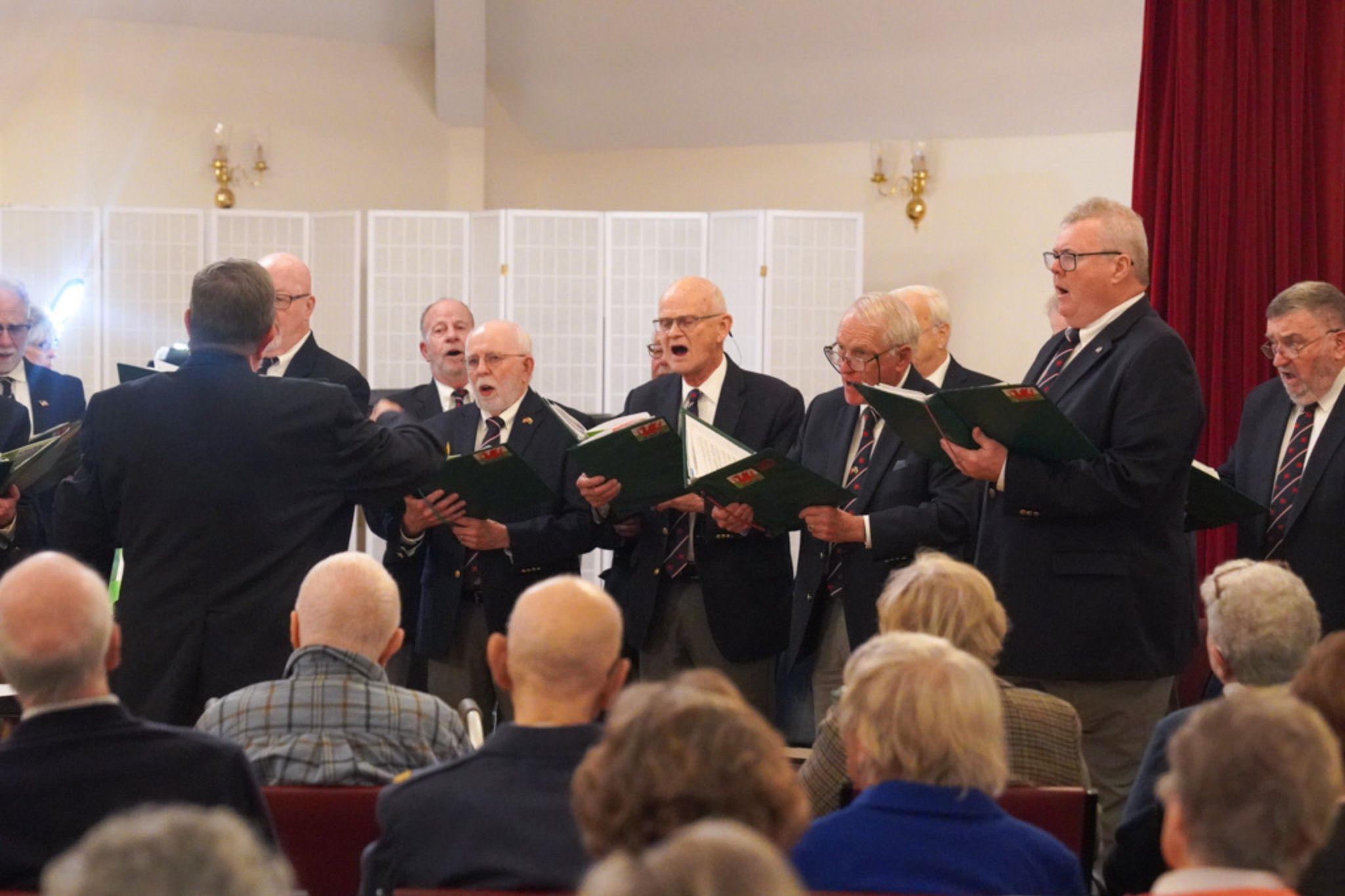 A choir of elderly men sings from sheet music on stage, directed by a conductor before an audience, their performance reflecting a lifetime of musical scholarship.