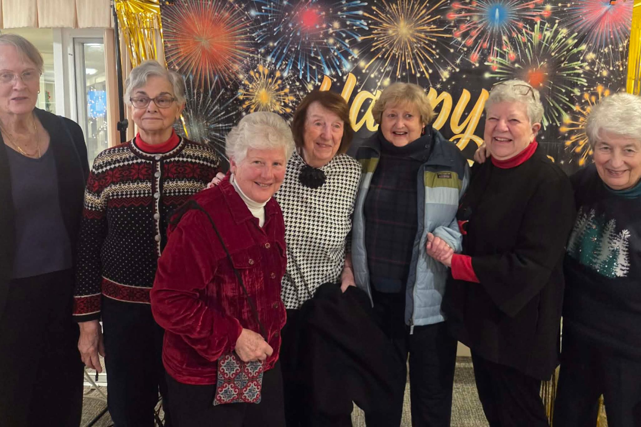Seven smiling older women stand together in front of a festive Happy fireworks-themed backdrop, capturing a joyful summer moment perfect for a photo essay.