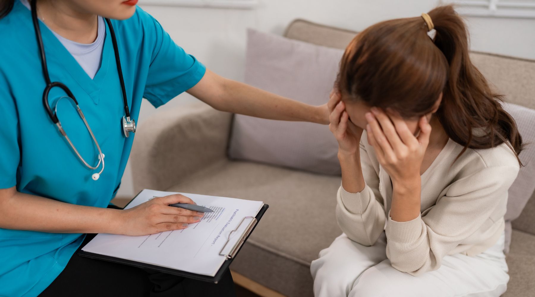 A nurse comforts a distressed woman on a couch, holding her shoulder and a clipboard with forms—offering support during a stressful holiday season.