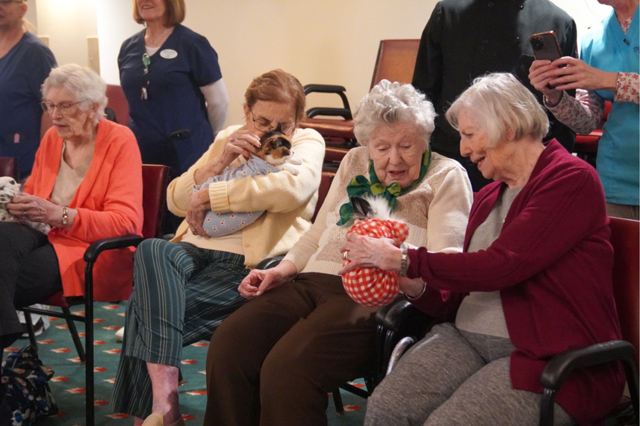 Four older women sit together, one holding a puppy while the others chat and share a wrapped gift, celebrating a recent scholarship achievement.