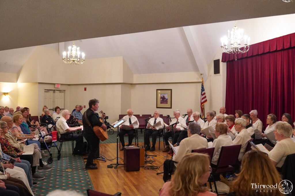 A man with a guitar leads a seated choir singing in a room filled with an audience, celebrating the power of music and scholarship.
