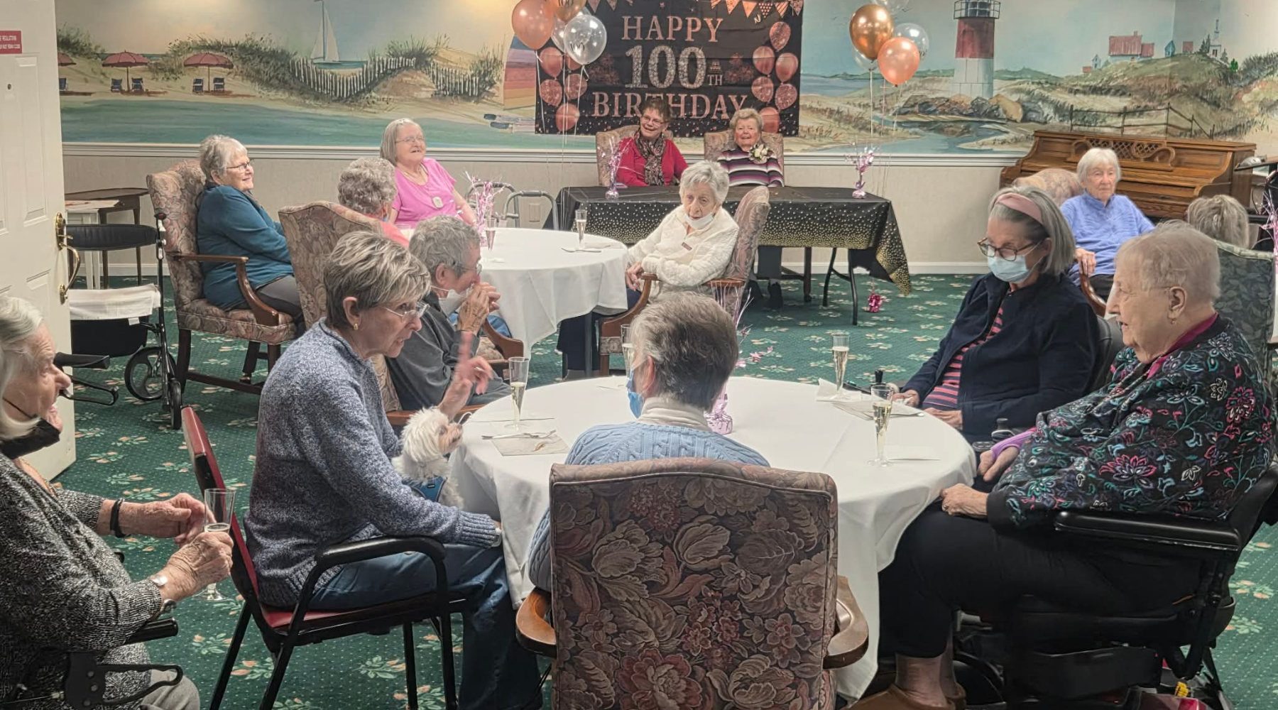 Elderly women sit around tables with a Happy 100th Birthday sign and balloons in the background, enjoying the celebration while staying cozy indoors this winter and taking care to avoid cold and flu season.