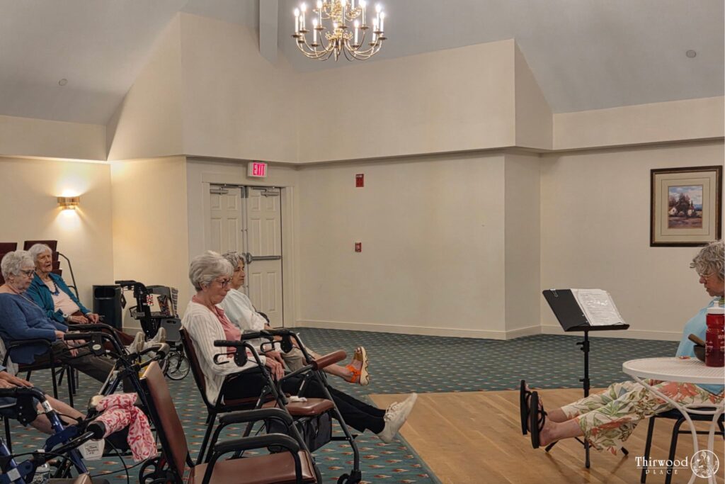 A group of elderly women sit in a room, some with walkers, participating in a seated exercise class as part of a scholarship program promoting wellness.