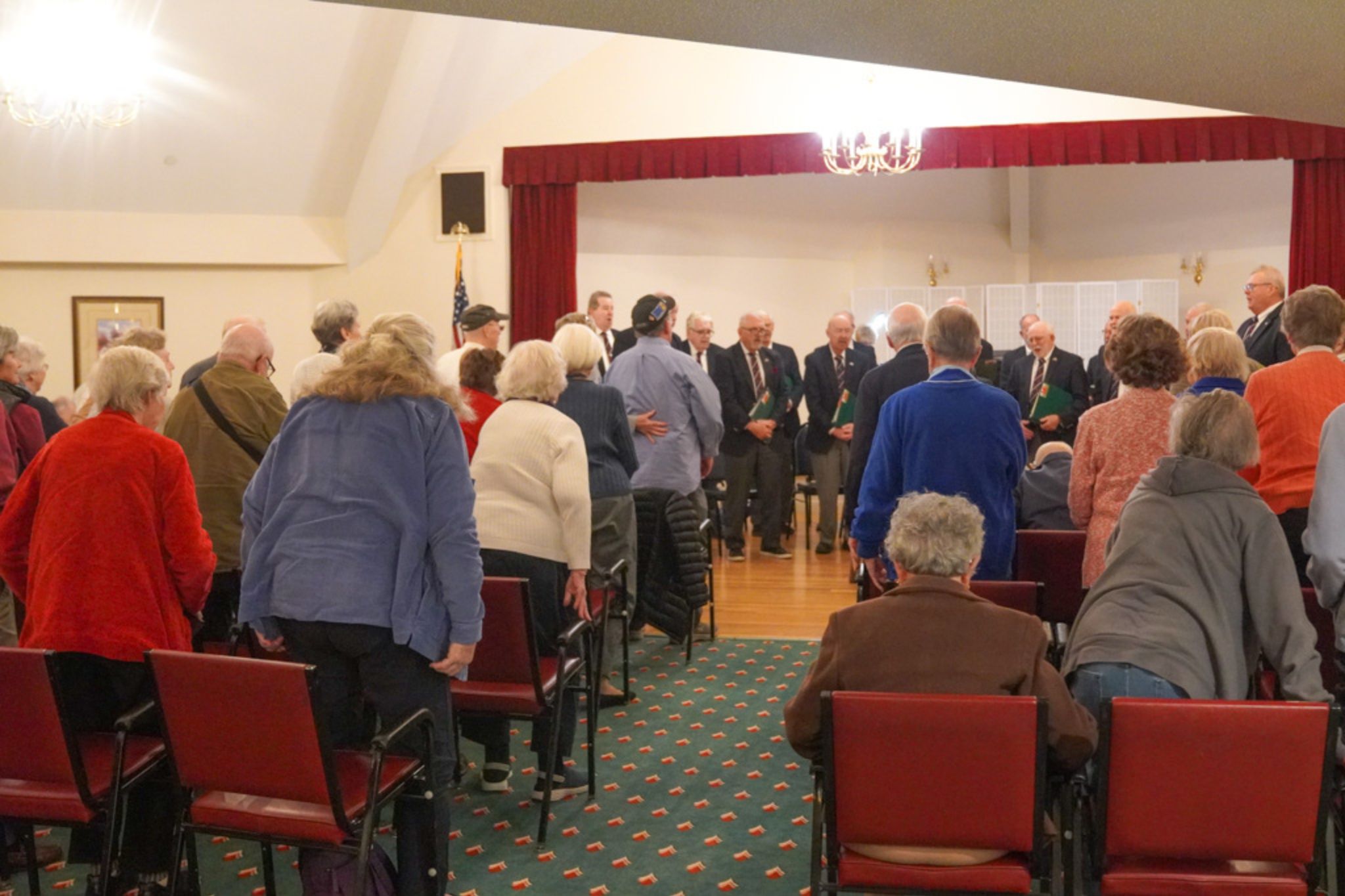 A group of elderly people stands and faces a small stage during an indoor scholarship event or gathering.