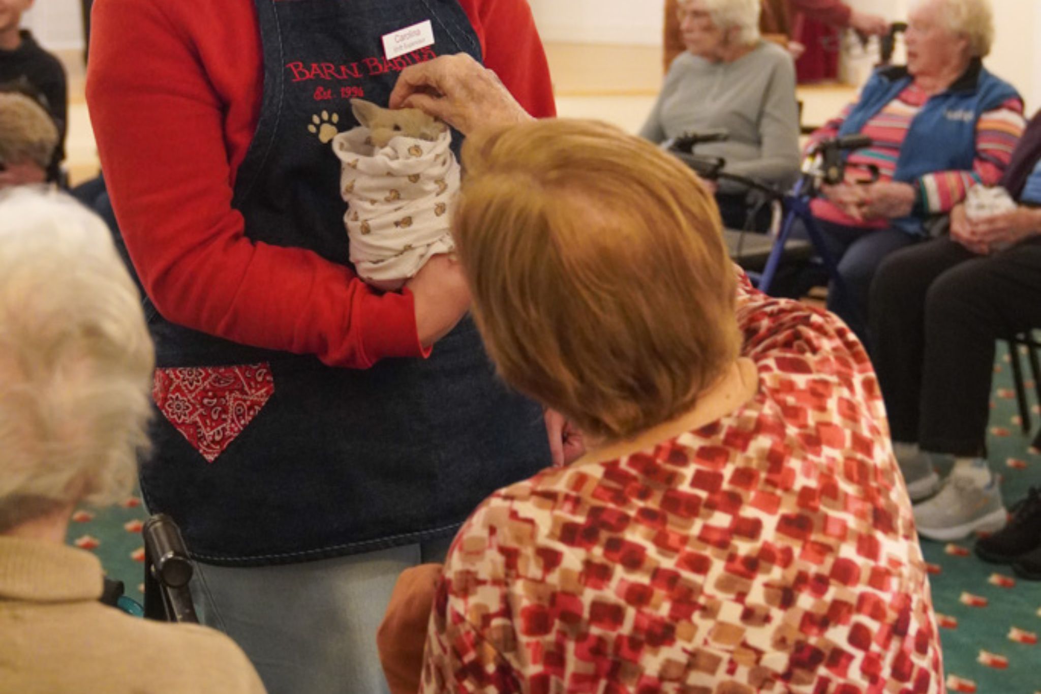A woman, visiting on a scholarship, shows a small animal to seniors seated in a group at a care facility or community center.