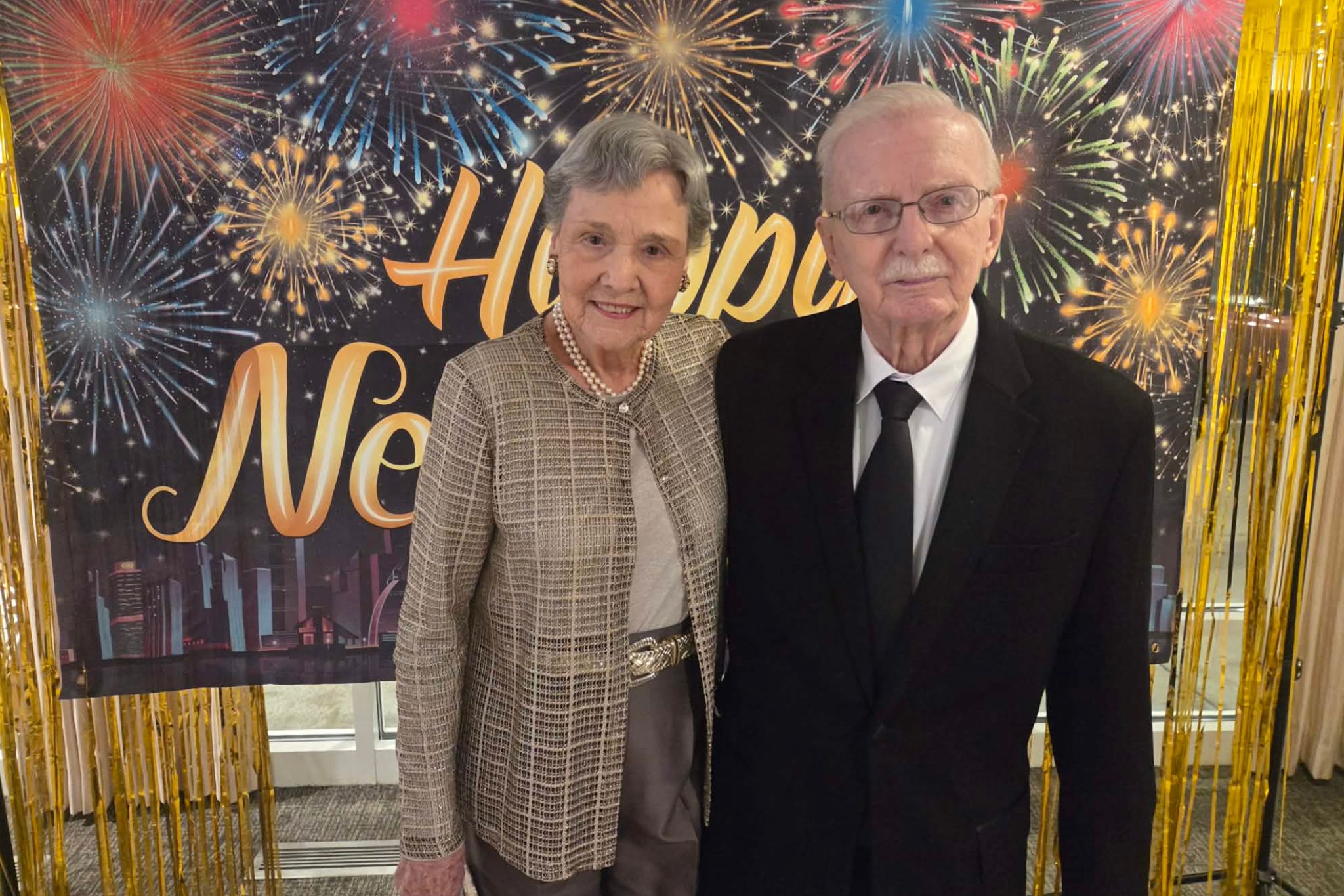 Elderly couple dressed formally standing in front of a Happy New Year fireworks backdrop, perfect for a photo essay capturing timeless celebrations beyond summer nights.