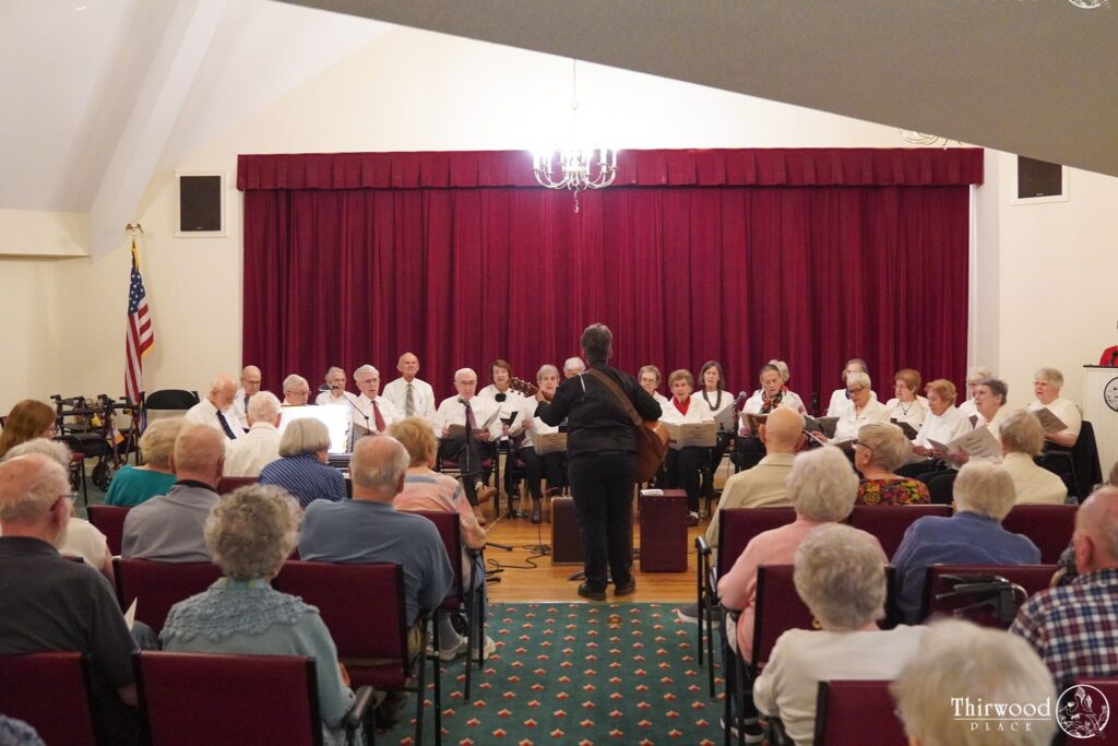 A choir performs on stage for an audience in a hall with red curtains and an American flag, celebrating recipients of a music scholarship.