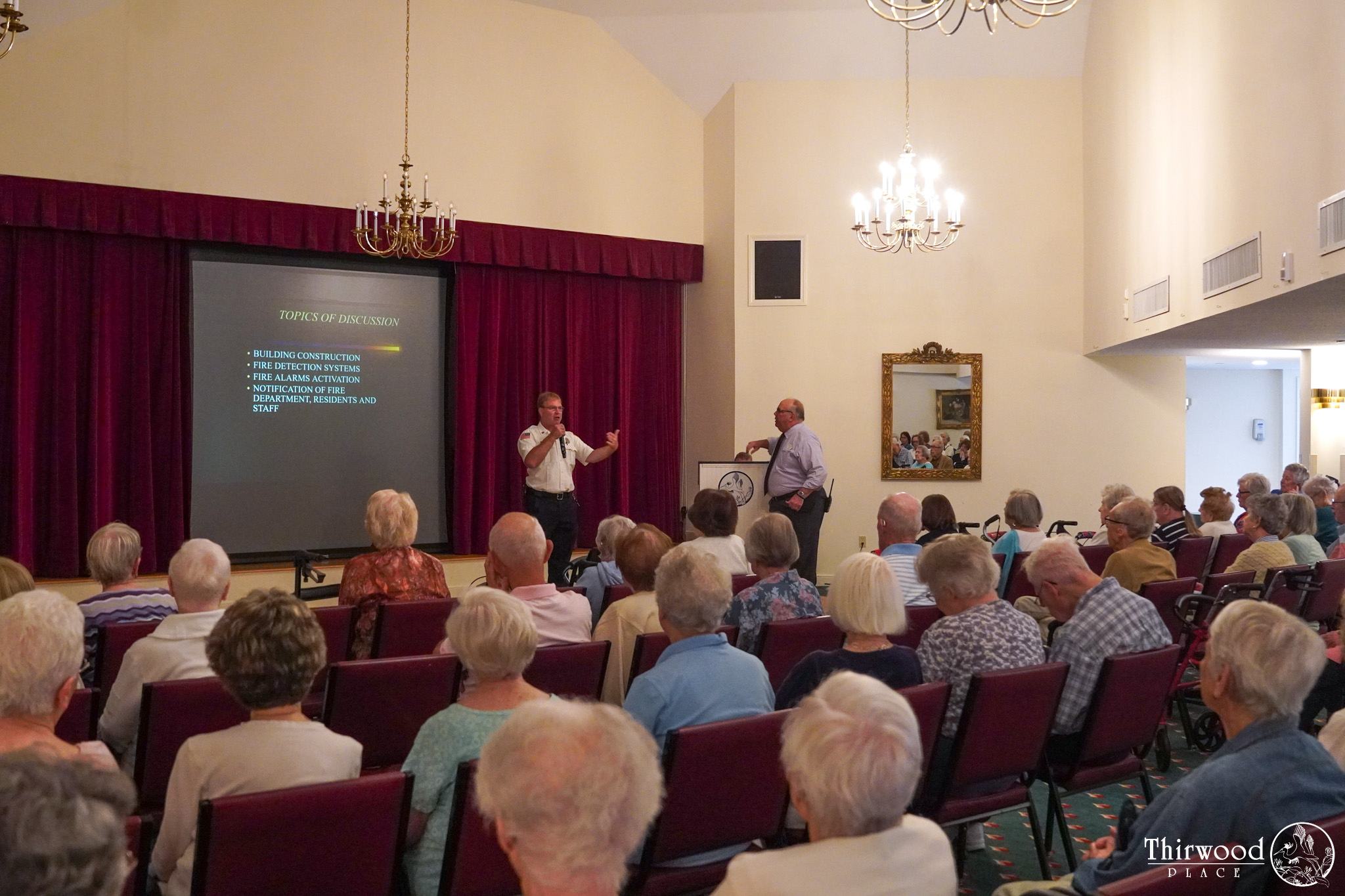 A speaker presents to a seated audience in a hall, with a projector screen displaying discussion topics on how to prevent cold and flu.