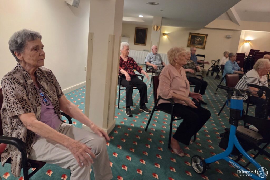 Elderly women sitting in chairs indoors, participating in a group activity or exercise session, share stories about their experiences and scholarship achievements.