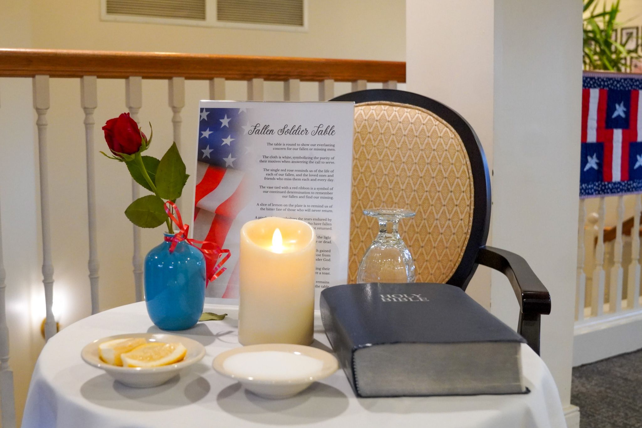A table with a candle, Bible, rose, glass, lemon slices, and a Fallen Soldier Table sign pays tribute and inspires scholarship in remembrance of those who served.