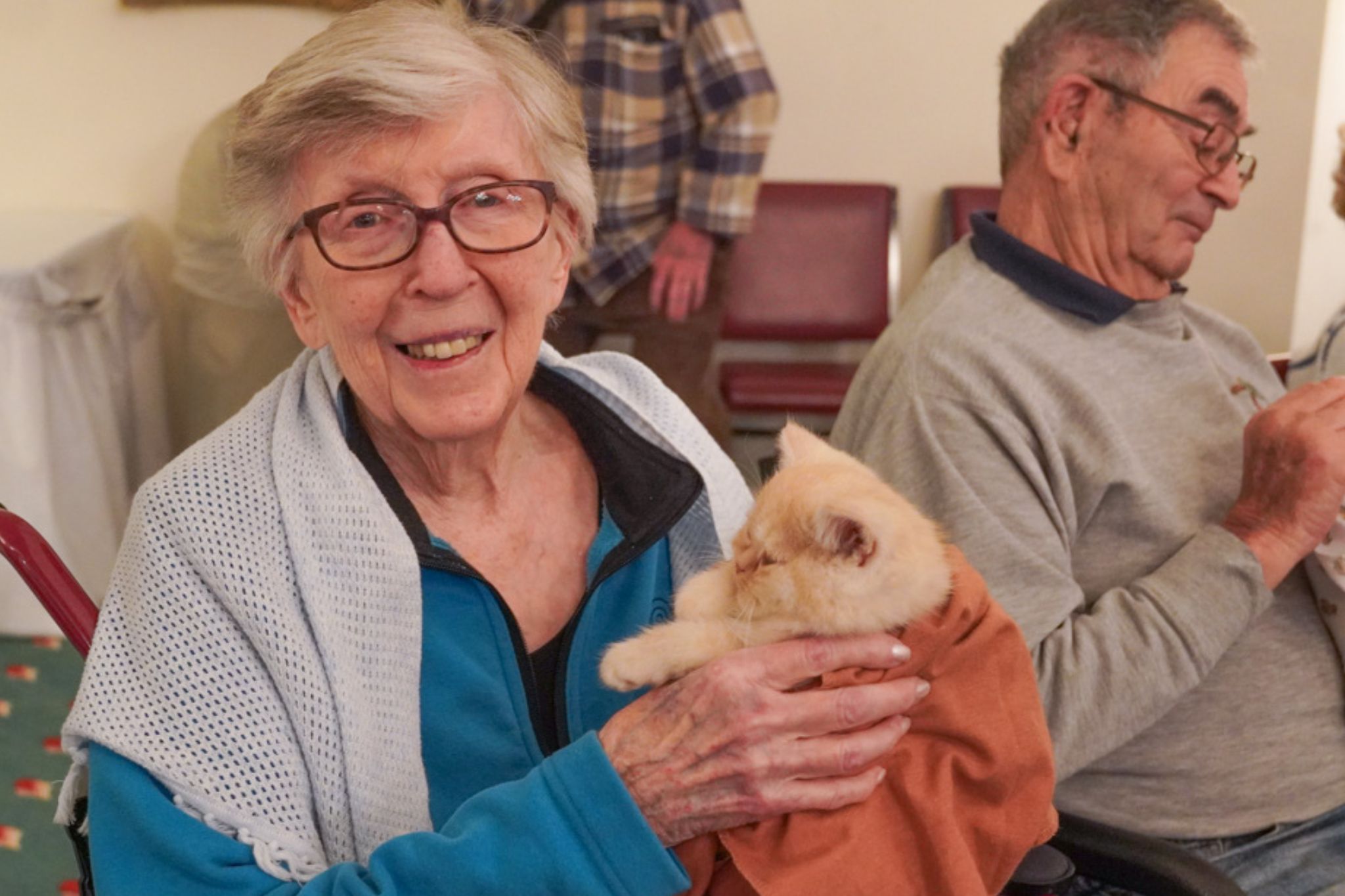 Smiling elderly woman holds a light-colored cat on her lap; man sits beside her, looking down, reflecting on her recent scholarship achievement.