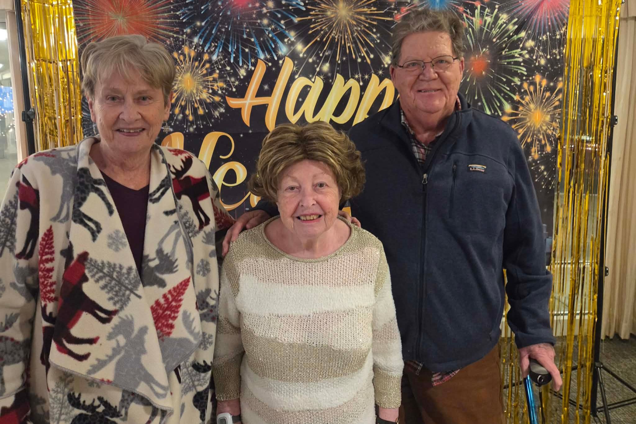 Three smiling older adults stand together in front of a Happy New Year fireworks backdrop, capturing a joyful moment perfect for a photo essay about celebration and togetherness.